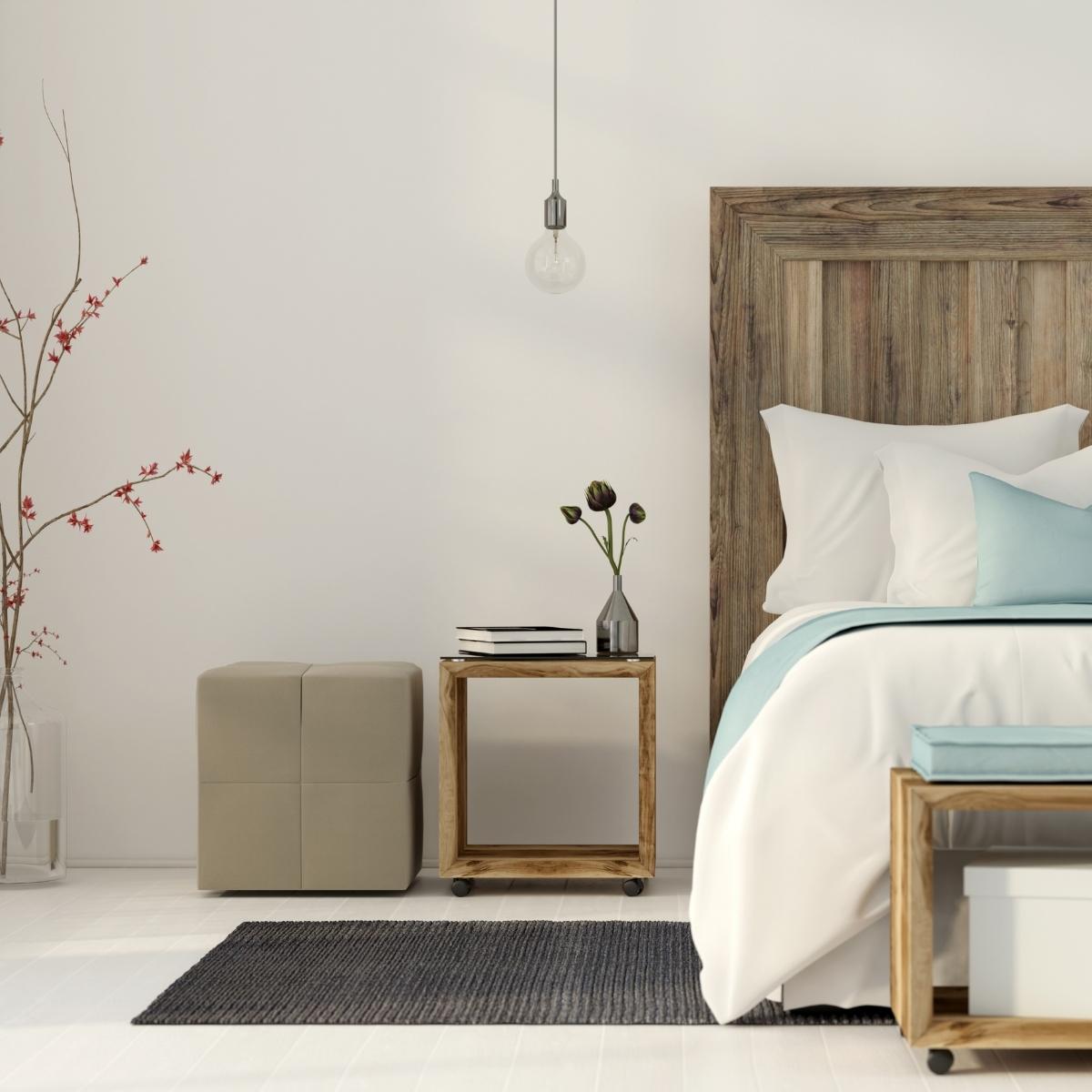 A modern bedroom designed for a good night sleep features a wooden bed with white and light blue bedding, a matching nightstand with books and a vase, a beige ottoman, a hanging light bulb, and a gray rug on white flooring.