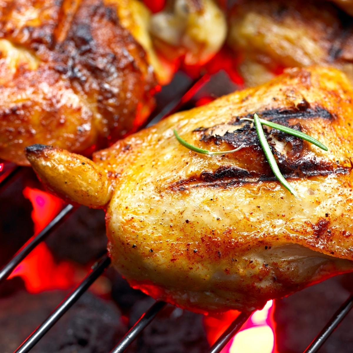 A close-up of a piece of grilled chicken with grill marks, seasoned and garnished with rosemary, sizzles on a metal grill over glowing coals—perfect for your garden summer-ready BBQ from Home Bargains.