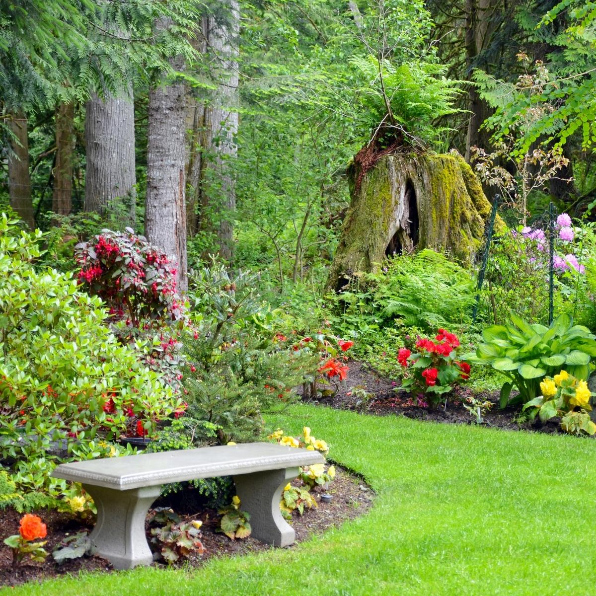 A concrete garden bench sits near colorful flowers, green plants, and shrubs in a landscaped garden bordered by tall trees and a mossy tree stump. The grass is neatly trimmed and lush—a serene spot worthy of a Sloane and Sons review.