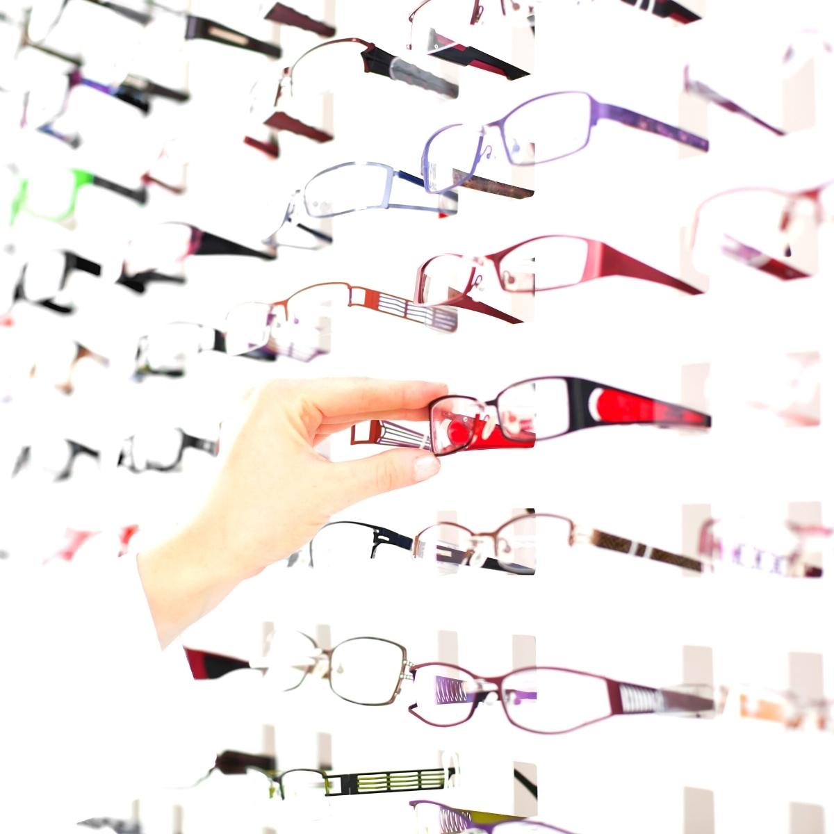 A hand reaches for a pair of red eyeglasses from a display of frames, highlighting the importance of glasses repair kits like SnapIt for keeping your eyewear in top shape.