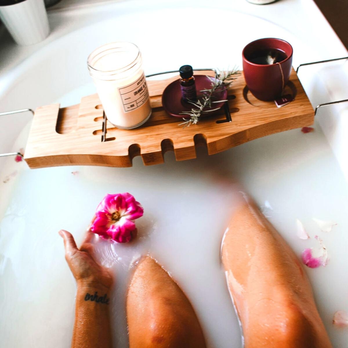 A person relaxing in a milky white bath, holding a pink flower. A wooden bath tray holds a lit candle, mug, small bottle, and rosemary—perfect for reading a Friction Free Shaving review or planning your next razor subscription. Pink petals float around.