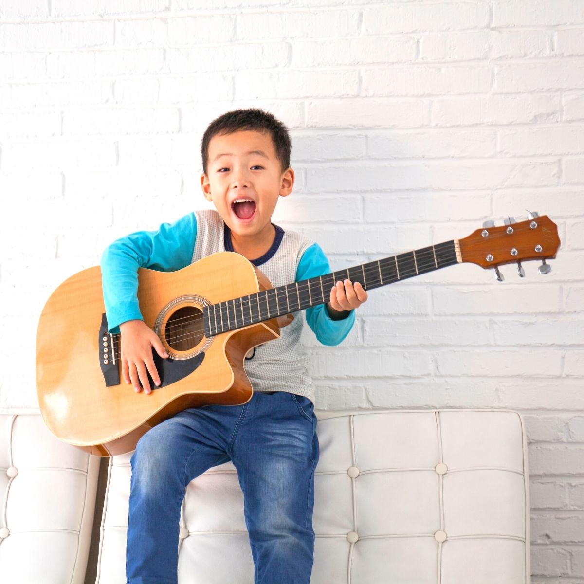 A young boy sitting on a white couch holds an acoustic guitar and appears to be singing or shouting, with his mouth open wide and an enthusiastic expression—a perfect example of fun and unique hobbies for children. The background is a white brick wall.