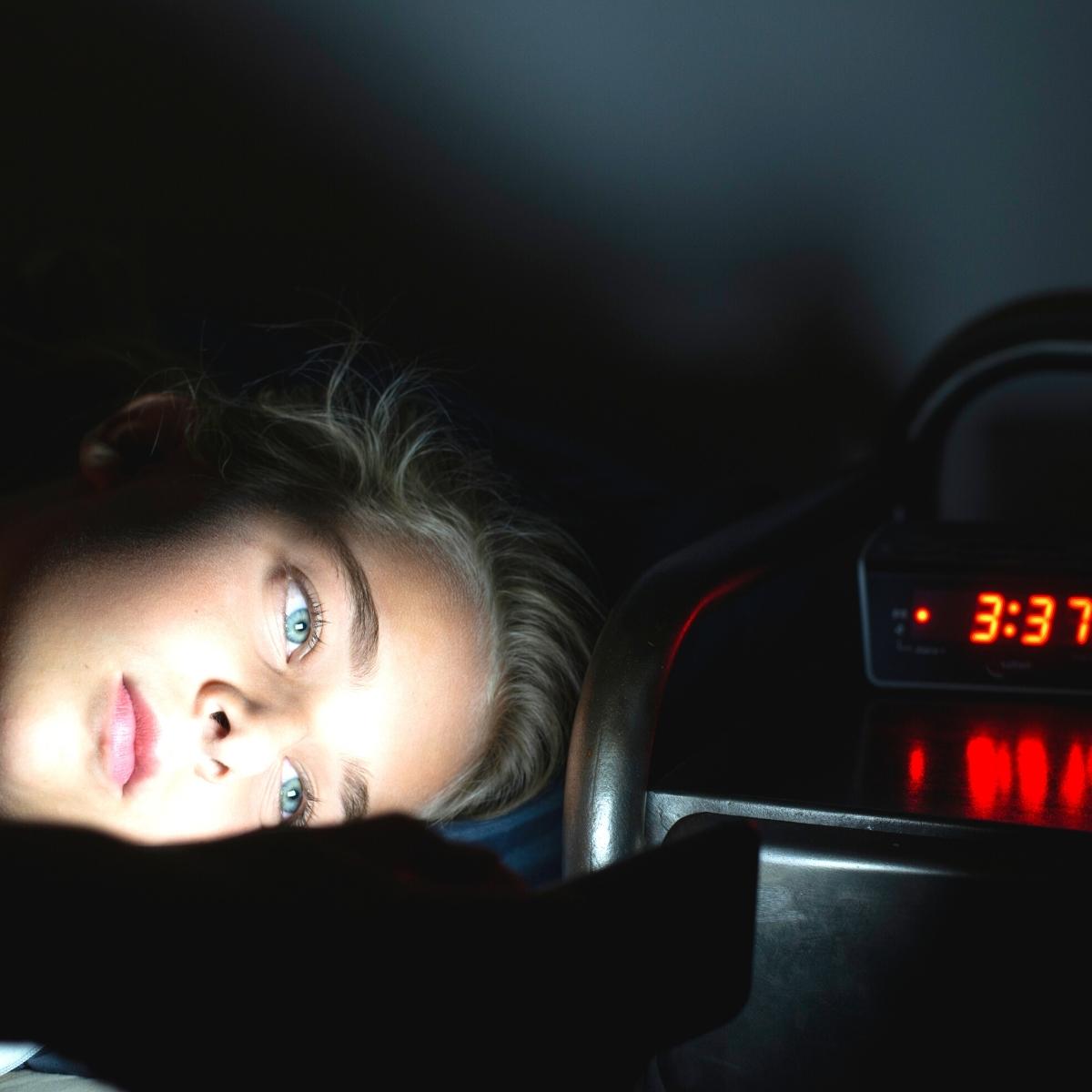 A person with blonde hair lies in bed in a dark room, illuminated by the light from a phone or tablet, perhaps searching for games to play when you can't sleep. An alarm clock on the nightstand shows the time as 3:37.