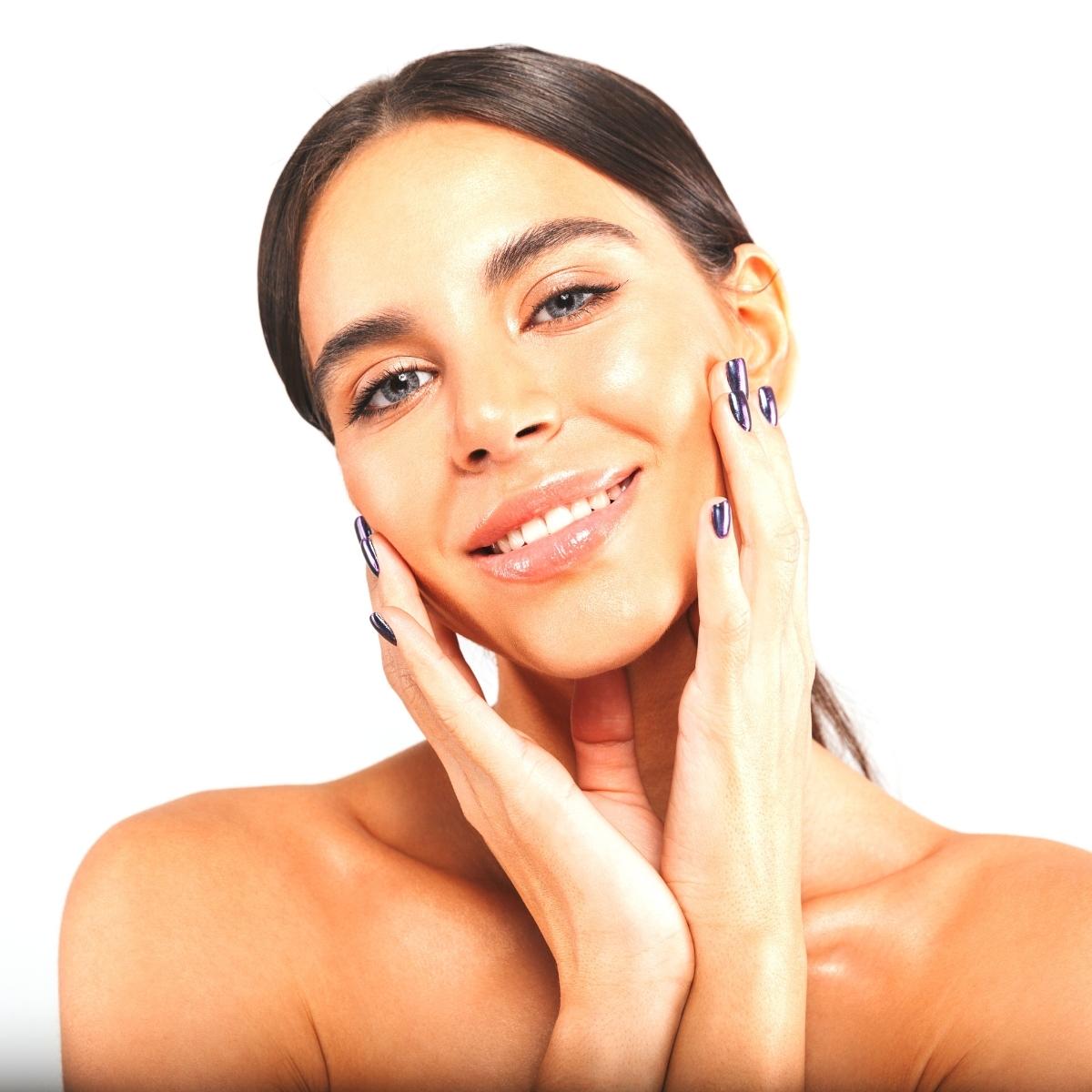 A woman with straight brown hair and glossy nails smiles at the camera, touching her face with both hands. She has smooth, glowing skin—showing no under eye dark circles—against a plain white background.
