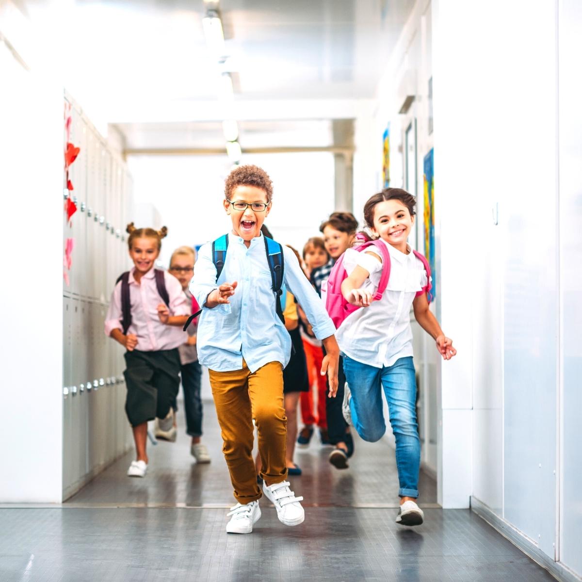 A group of children with backpacks run down a brightly lit school hallway, smiling and appearing excited—showcasing the joy that strong parent engagement brings to a child’s education, with lockers and classroom doors visible on either side.