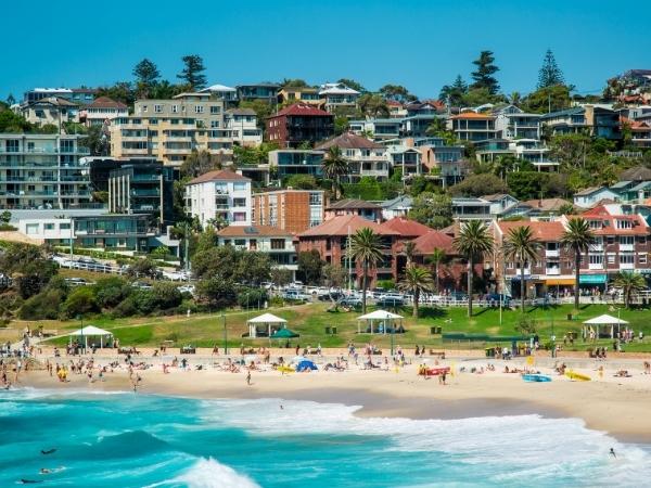 A sandy beach with people swimming and sunbathing near the shoreline, grassy areas, palm trees, and a variety of residential and commercial buildings on a hillside in the background—perfect for exploring Halton’s scenic Waterfront Trail.