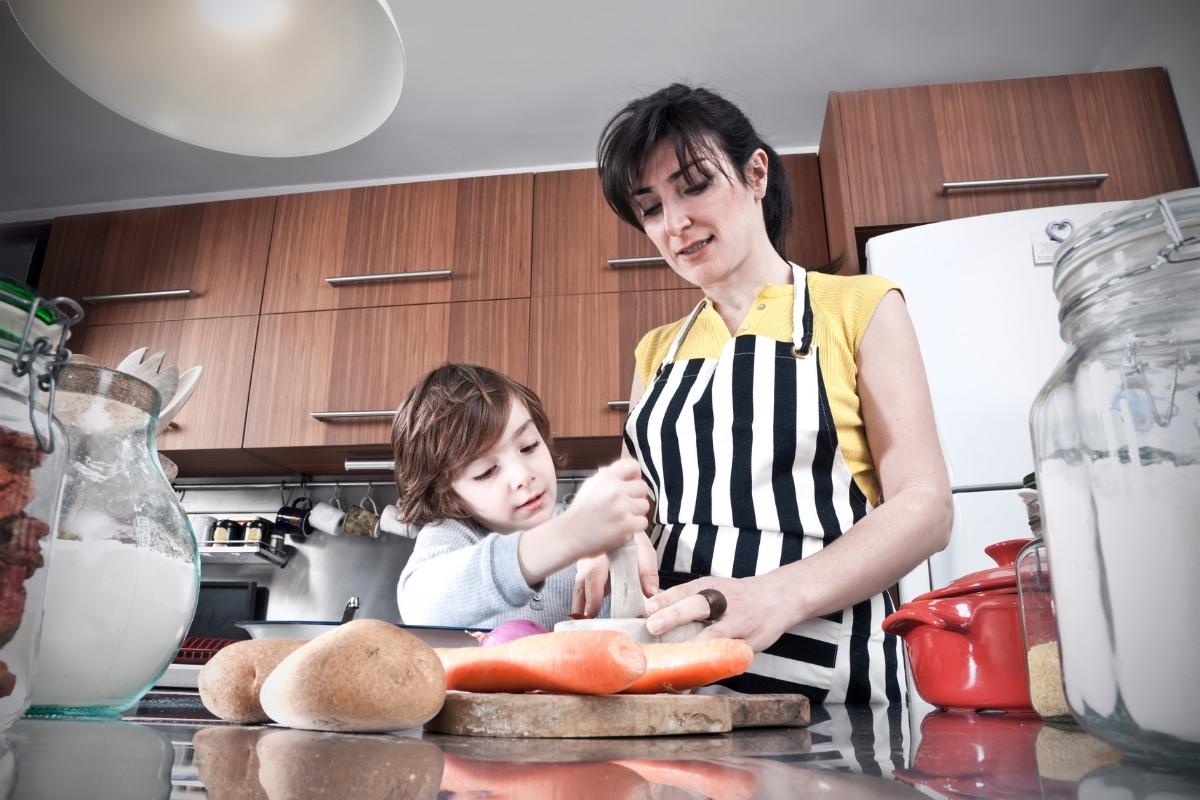A woman and a child stand in a kitchen preparing food together. The woman wears a striped apron as the compassionate child helps with mixing—an everyday moment of parenting that nurtures kindness and connection.