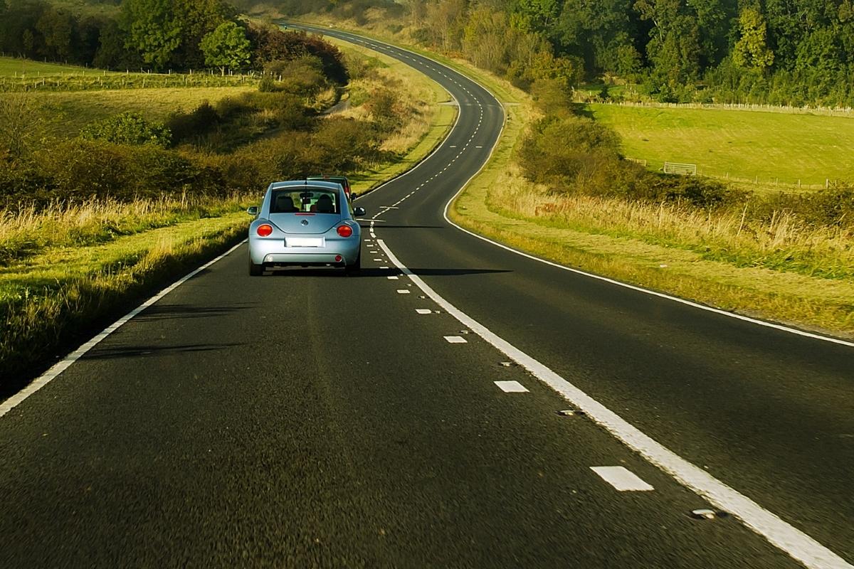 A light blue car drives along a winding two-lane rural road, surrounded by green fields and trees under a clear sky. In the wake of Covid19, owning a car offers a peaceful countryside escape and personal freedom amidst current events.