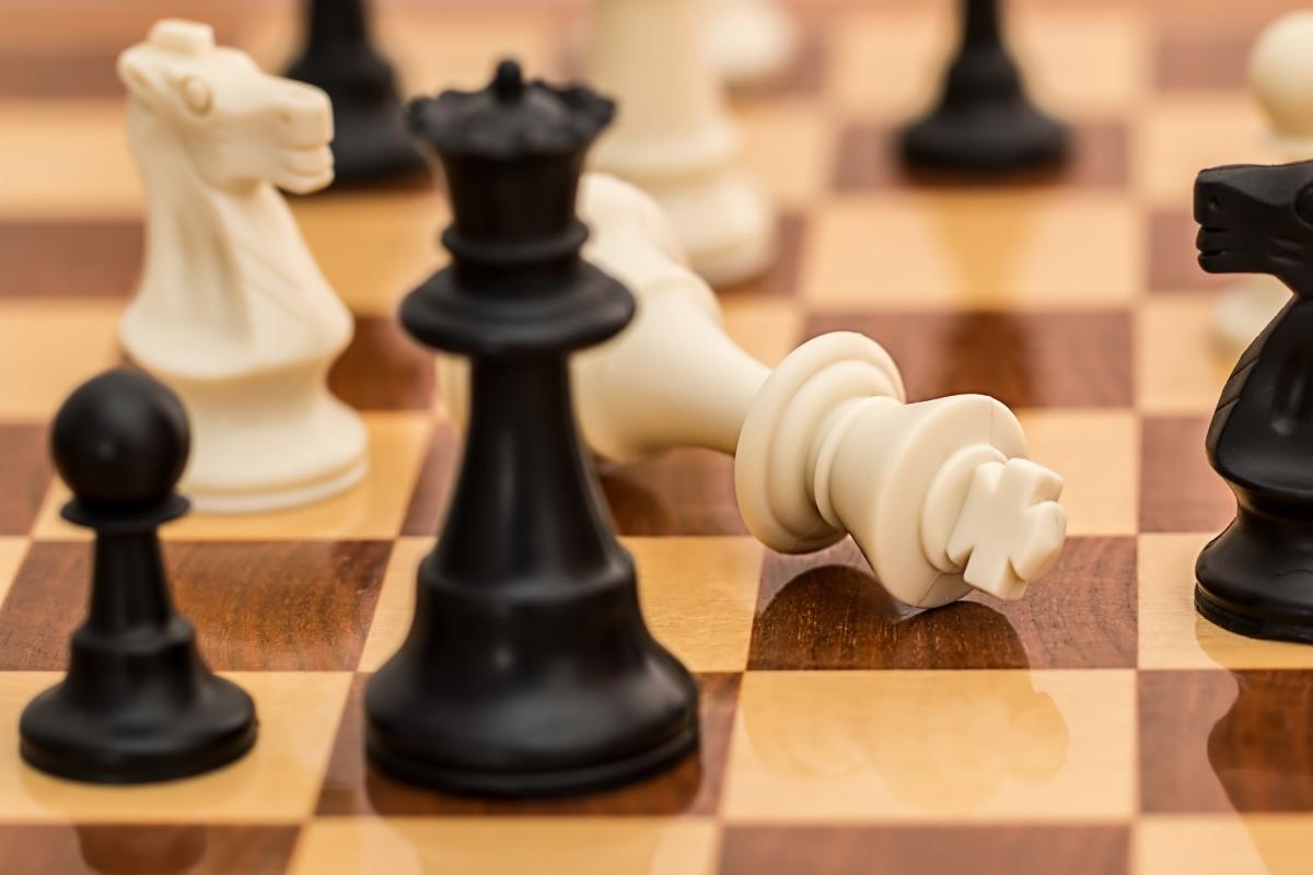 A close-up of a chessboard shows several pieces, including a toppled white king lying on its side, as if in sleep, surrounded by standing black and white chess pieces. The board features alternating light and dark squares.
