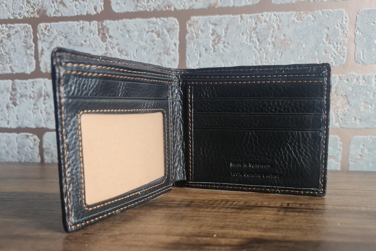 A black leather bifold wallet with tan stitching, displayed open on a wooden surface in front of a light-colored brick wall. Featuring card slots and a transparent ID window, it’s a stylish pick for men's gifts or thoughtful gifts under £40.