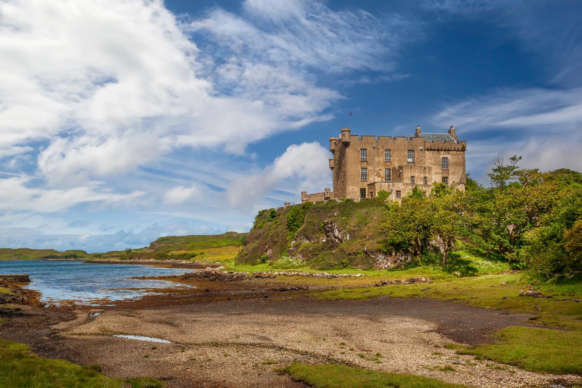 A stone castle sits on a grassy hill overlooking a calm body of water, with trees nearby and a partly cloudy blue sky above, offering a picturesque setting that highlights the importance of history lessons in schools.