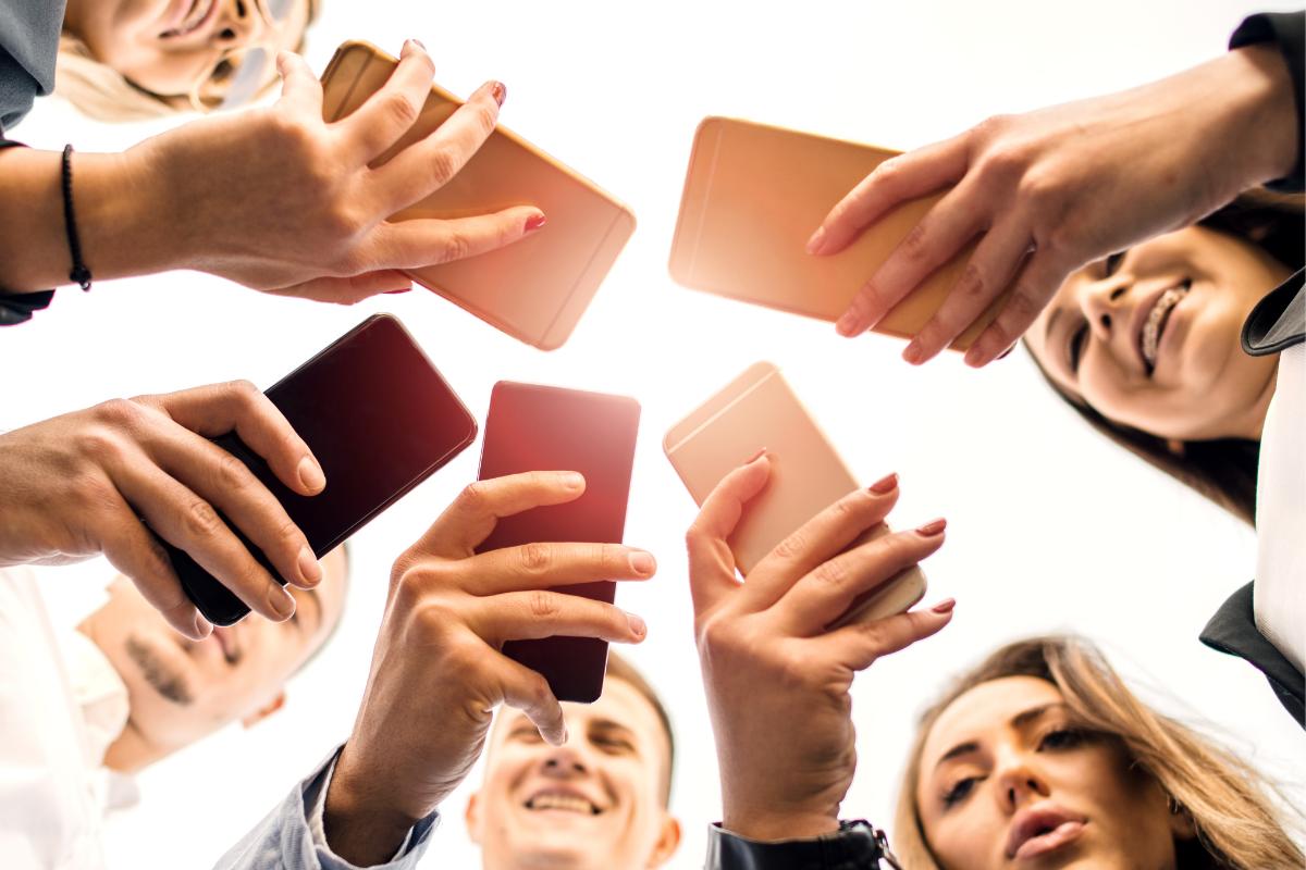 A group of people stands in a circle, holding smartphones above their heads and looking up—captured from below with faces partly visible and bright light behind them—symbolizing teaching children social media and child online safety.