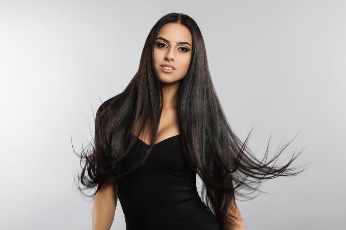 A woman with long, straight black hair wearing a sleeveless black dress stands against a plain light gray background, looking at the camera with a slight smile. Like many women with long hair, her flowing locks are spread out, suggesting movement.