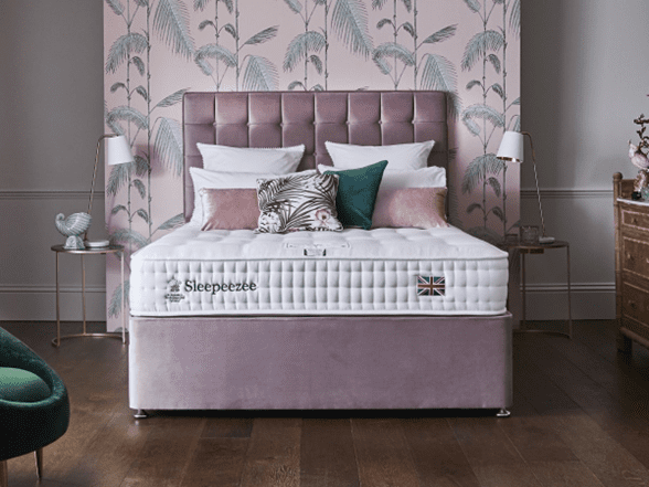 A neatly made bed with a tufted pink headboard, white and patterned pillows, and a luxurious mattress. The room features pink palm leaf wallpaper, two side tables with lamps, and a wooden floor.