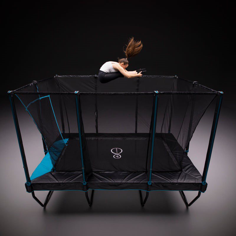 A person with long hair jumps and tucks their legs on a rectangular trampoline with safety nets, capturing a moment of childhood bliss, viewed from above in a studio setting against a dark background.