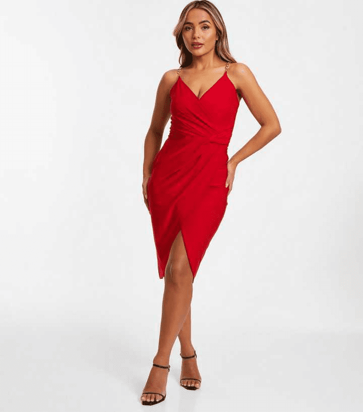 A woman stands against a plain background wearing a sleeveless, knee-length red dress with a wrap-style front and thin straps—perfect dressing for Valentine’s Day. She completes the look with black strappy high-heeled sandals.