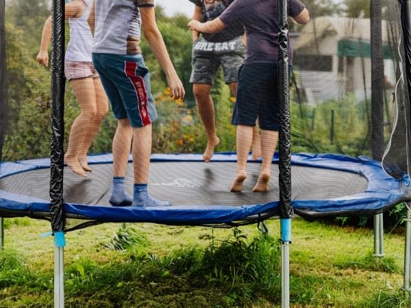 Four kids are jumping together on a trampoline outdoors, surrounded by grassy plants and trees—perfect for sparking a love for nature. A safety net keeps them secure in this parent's guide–approved summer scene.