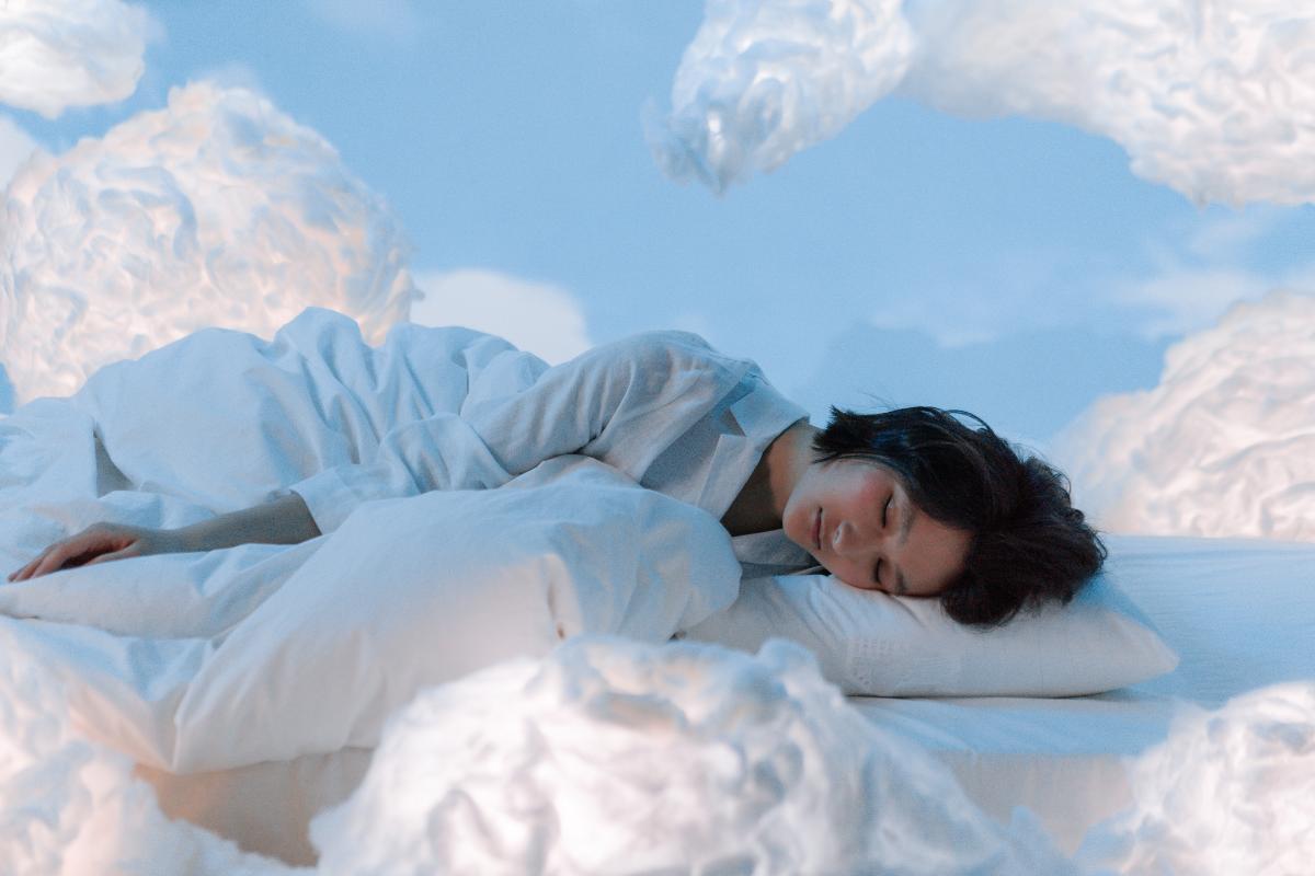 A person is lying on a bed with white bedding, resting their head on a pillow. Surrounded by fluffy, cloud-like formations against a blue sky, they appear lost in dreams, creating a serene and dreamy atmosphere.