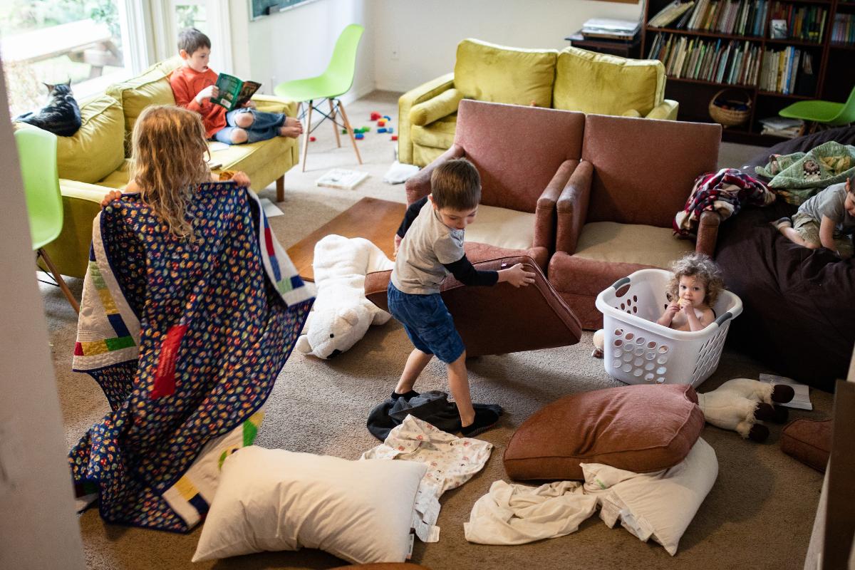 Four children play in a living room scattered with pillows, blankets, and toys. One stands with a quilt, another pushes a laundry basket with a child inside, and another reads on the couch—perfect for those organizing with children moments.