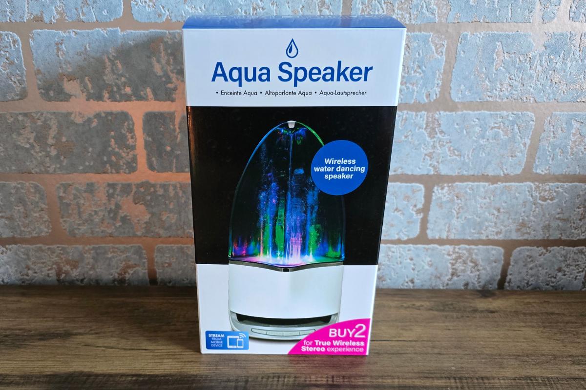 A boxed Aqua Bluetooth Speaker sits on a wooden surface in front of a brick wall. The packaging, from Wicked Uncle, highlights the speaker’s colorful lights, water feature, wireless operation, and stereo pairing capabilities.