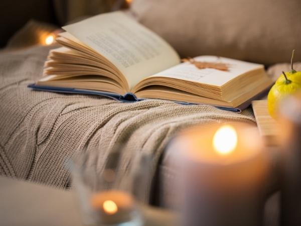 An open book rests on a beige knitted blanket with a lit candle and an apple nearby; the scene appears perfect for enjoying the best cosy crime books in a softly lit, inviting atmosphere.