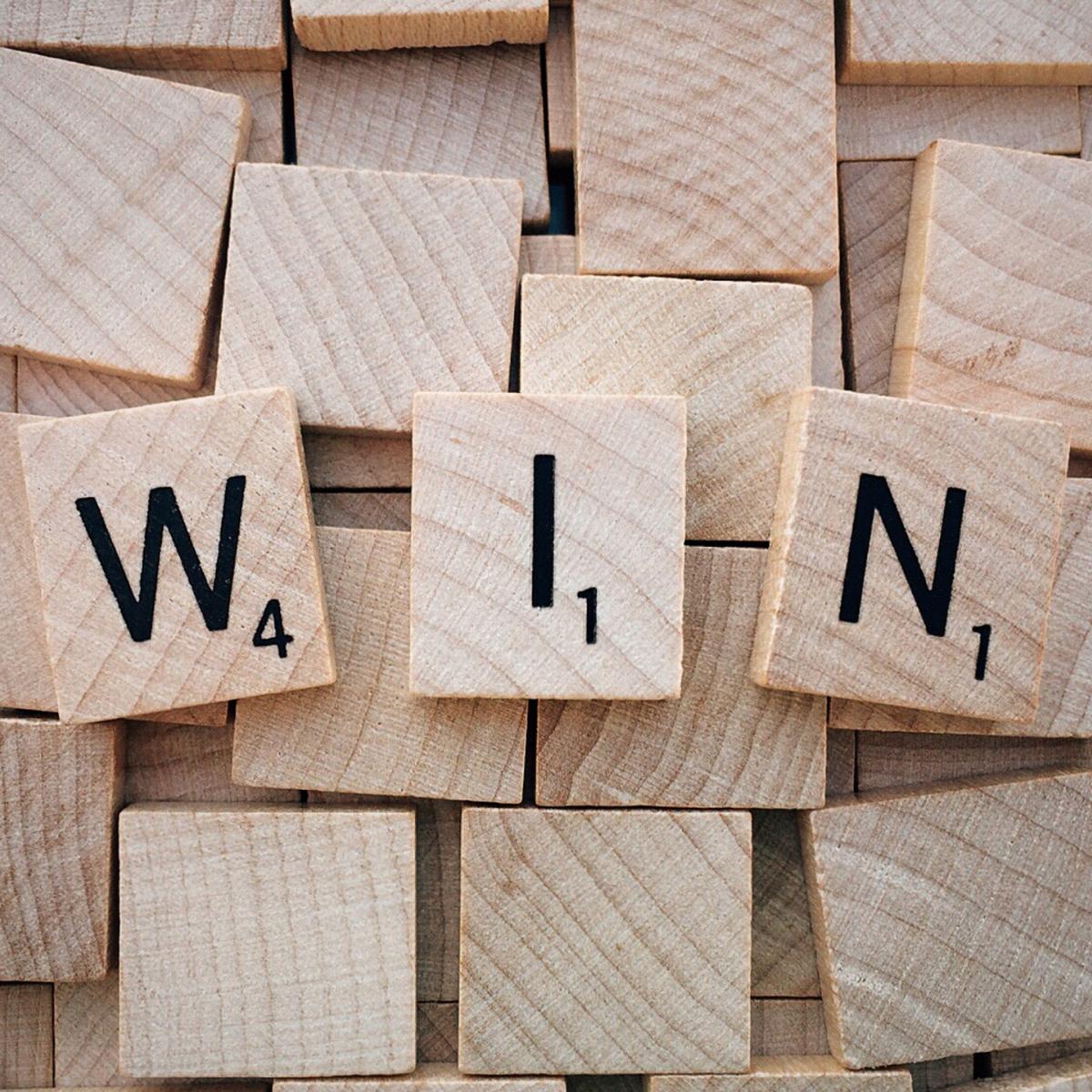 Three Scrabble tiles spell the word "WIN" against a background of overlapping, blank wooden tiles—reminding you to enter to win a book in our exciting book giveaway. The letter values are W (4), I (1), and N (1).