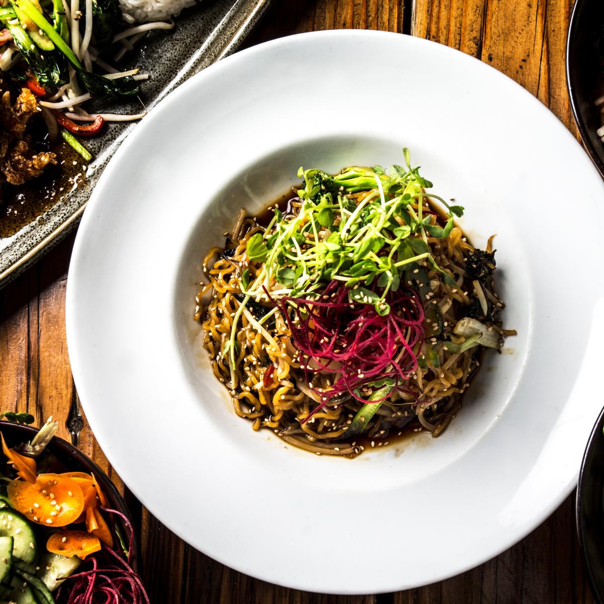 A white plate of noodles topped with thinly sliced vegetables and microgreens sits on a wooden table, surrounded by vibrant dishes—perfect for practicing your dining etiquette while enjoying a colorful meal.