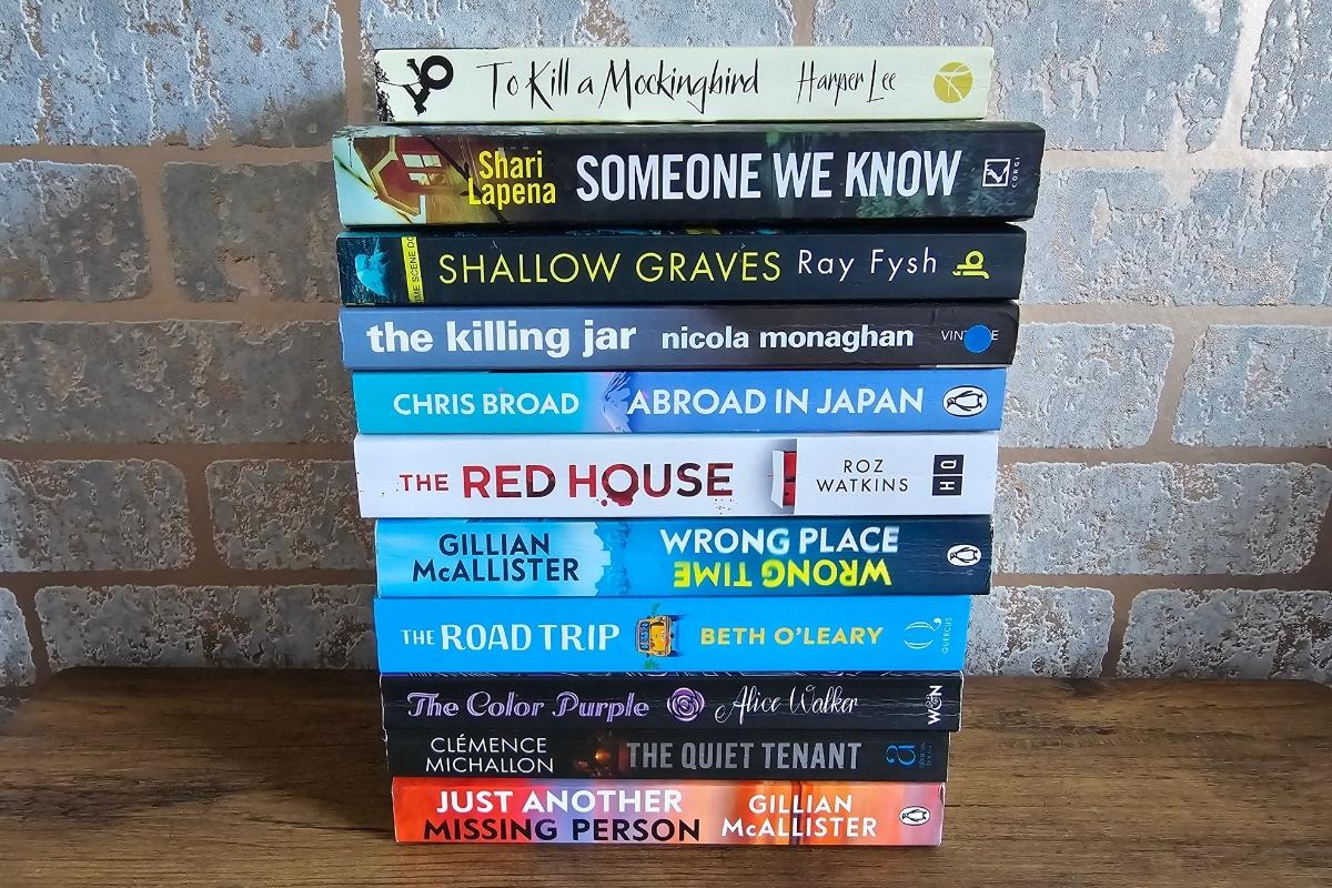 A stack of ten books sits on a wooden surface against a brick wall—perfect inspiration to try the best apps to track books and keep your own reading tracker up to date as you work through each compelling title.